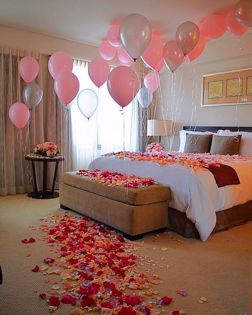 اتاق خواب عروس و دامادی که کف آن با گلبرگ های صورتی، قرمز و زرد و سقف آن با بادکنک های هلیومی صورتی رنگ دکور شده است